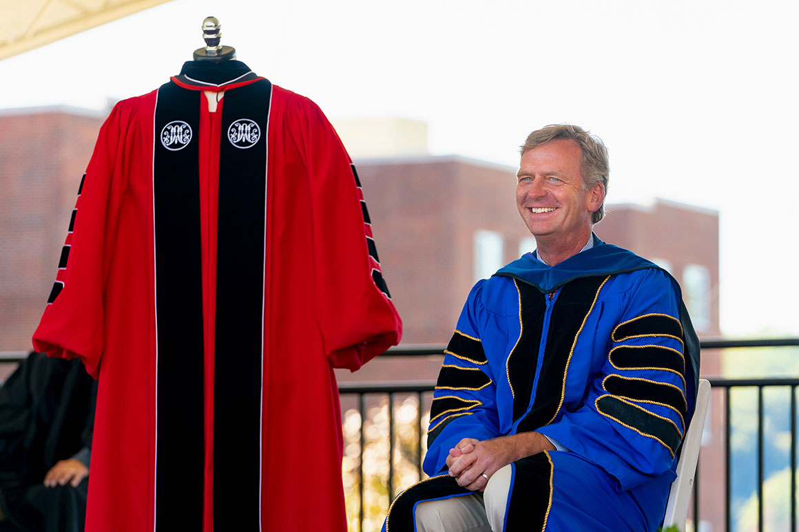 Kevin Weinman sitting onstage wearing the robe associated with his earned degree next to the Marist red robe