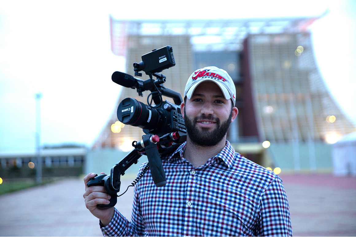 Image of Alec Rizzo standing outside of Jubilee House in Accra, Ghana. He has a video camera over his right shoulder and is wearing a Marist baseball cap