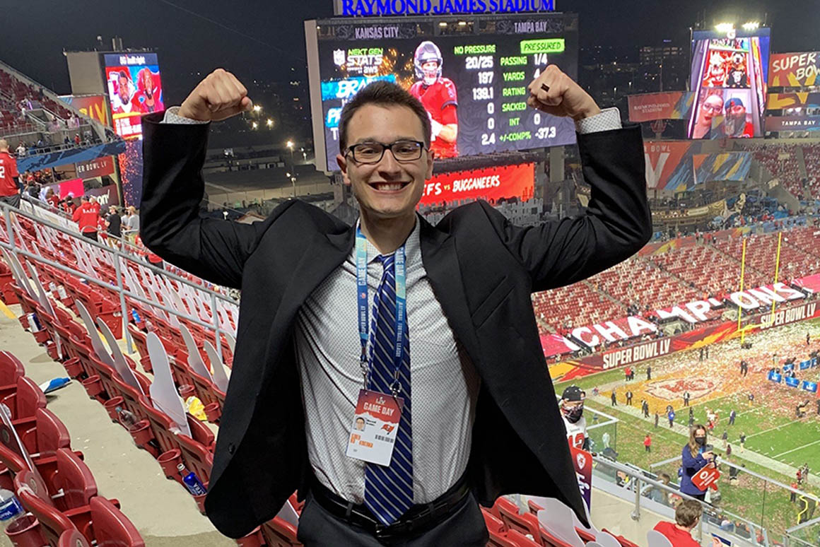 Image of Maxwell standing in the Raymond James Football Stadium with his arms raised above his head in a show of strength. He's wearing a grey suit, blue and white tie, and press credentials